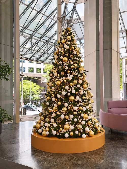 4m Christmas tree standing on a gold base decorated in gold and white baubles with a large star. The tree stands in the corner of a large city foyer in the window.