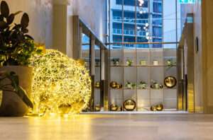 Large LED baubles glow in the foreground and oversized baubles sit in a wooden shelf between two elevators in a city building foyer.