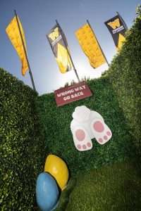 An Easter hedge maze in a garden with sign reading "Wrong way go back" and below it the rear end of a rabbit disappearing through the hedge. There is also a blue and a yellow easter egg on the ground.