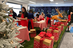 A group of adults busily decorating Gingerbread Houses at tables with red checkered tablecloths. Red and brown wrapped presents line the right hand side. In the background, a snowy backdrop of trees and a large reindeer.
