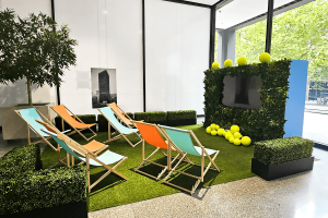 A screening area to show off the tennis matches for the Australian open. Beach chairs are spread-out on fake grass. The chairs are all facing towards a hedge wall with a tv in bedded in the middle.