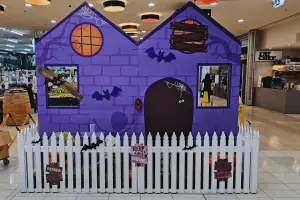 A purple haunted house is surrounded by a white picket fence. It stands in a shopping centre.