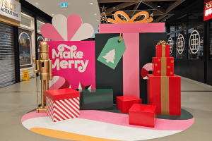 A shopping centre photo zone with Large pink and orange 2D gifts. They read Make Merry. In front of them stand a gold nutcracker and 3D gifts.