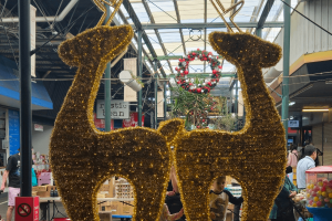 Large Golden tinsel and LED reindeers stand back to back. Through them we see a market scene, with red, white and black checkered wreaths.