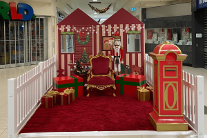 A deep red carpet and house backdrop make up a Santa scene in a shopping centre. There are large gift props and red velvet throne. In the foreground is a red and gold mailbox.