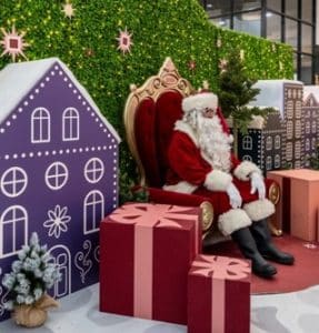 Santa sits on a red and gold throne in front of a lit hedge wall and mini gingerbread houses.