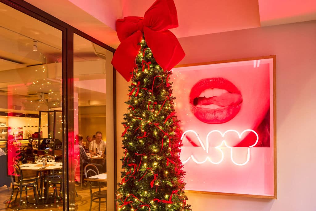 Large Christmas red velvet bow sitting upon a cone style Christmas tree, in front of a neon BABY sign at a restaurant entrance
