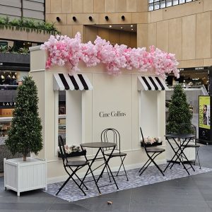 Box-like display with a window and black/white awning and cherry blossom overhead. Black cafe-style seating is in front of the display atop a stone decal