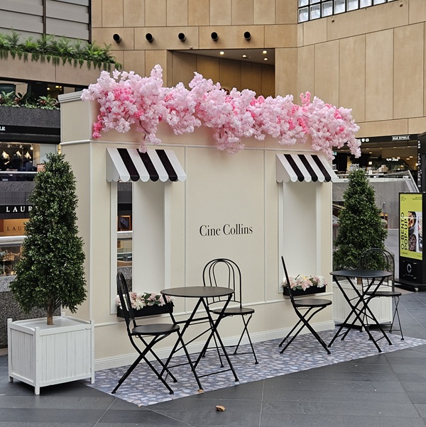 Box-like display with a window and black/white awning and cherry blossom overhead. Black cafe-style seating is in front of the display atop a stone decal