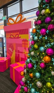 Coloruful pink, orange and yellow Christmas photobackdrop in the shape of a giftbox next to a colourful Christmas tree on pink base.