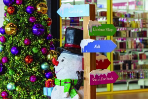 Commercial Christmas decoration in bright colours featuring a snowman and a Christmas tree, positioned next to a wayfinder sign pointing to the North Pole and various retail shops of a shopping centre precinct.