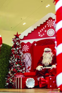 Santa sitting in front of a candy-themed house next to a candy decorated christmas tree.