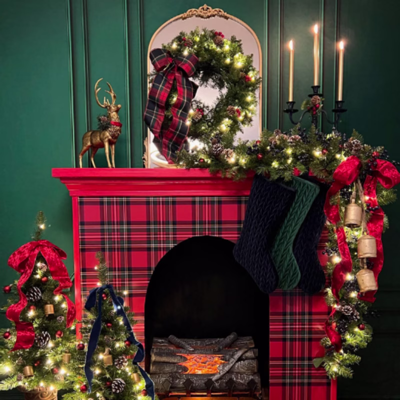 A tartan covered fireplace decorated with a traditional Christmas garland with lights and red bow. Green stocking hang from the fireplace and a Gold reindeer sits on top. 3 candles are alight on the fireplace. 2 Small decorated Christmas trees sit by the fire.