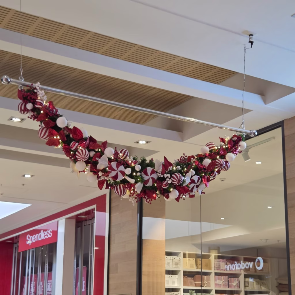 A candy-themed festive decor display featuring peppermint candy ornaments, baubles, and fairy lights suspended from a long silver metal frame in a shopping centre.