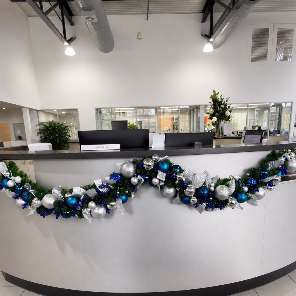 A christmas garland with silver and varying shades of blue baubles, and silver leaves with blue berry picks. The garland is hanging underneath a desk.