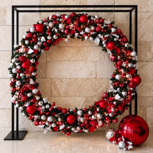 An oversized Christmas wreath adorned with red and silver baubles and metallic leaves, suspended within a sleek black square metal frame against a textured stone wall as a grand commercial festive decor display.