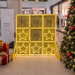 Interactive Christmas shopping centre display with a large 3×3 tic-tac-toe game set within a gold frame, decorated with gold stars and white snowflakes,alongside red and green control buttons at the front.